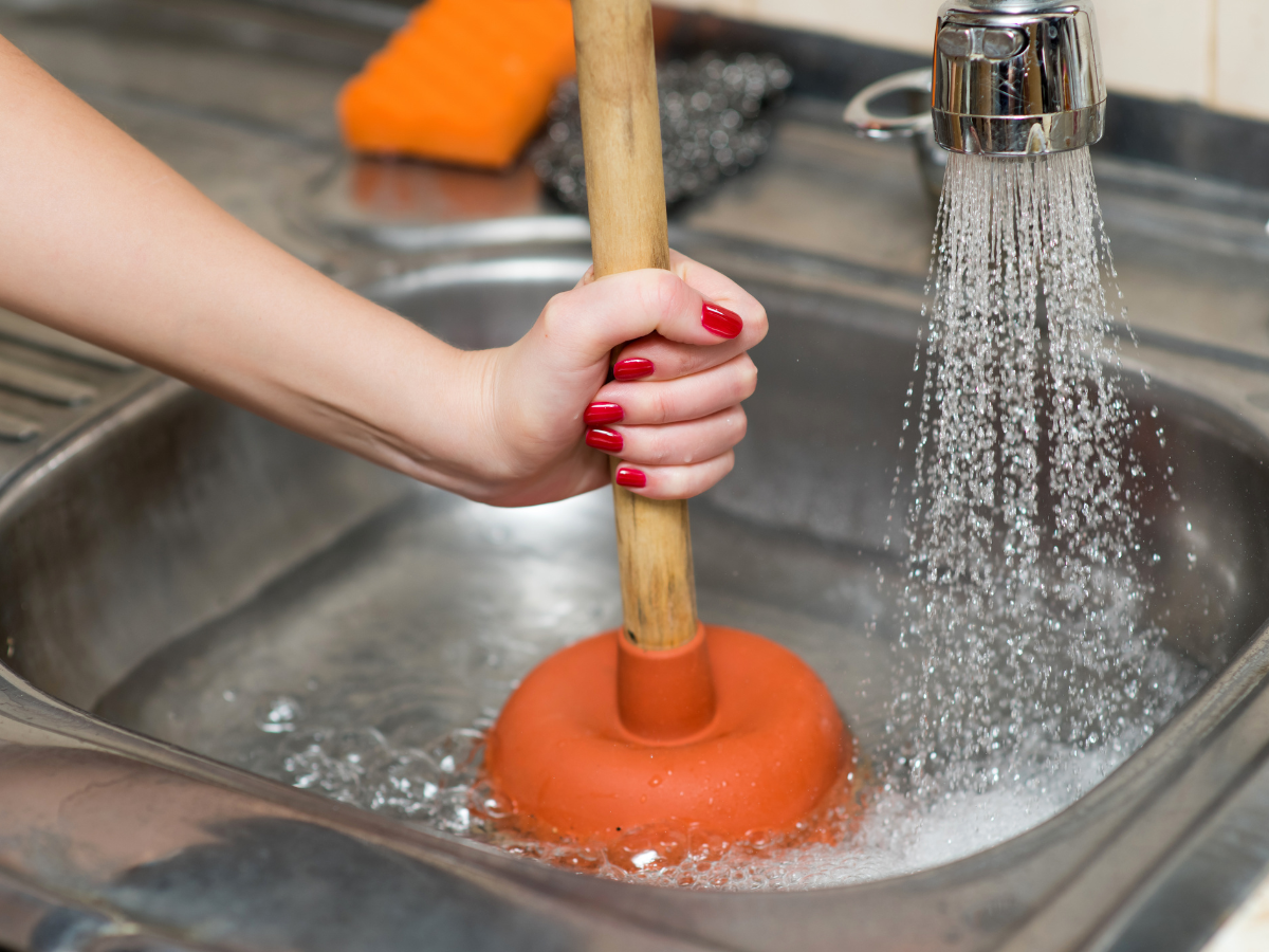 A person uses a plunger in a kitchen sink filled with water, while the faucet is running, focusing on preventing clogs and ensuring smooth drainage.