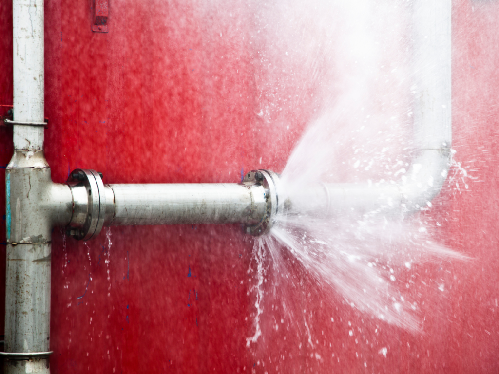 A high pressure metal pipe with a large leak sprays water against a red wall, causing water to flow in multiple directions.