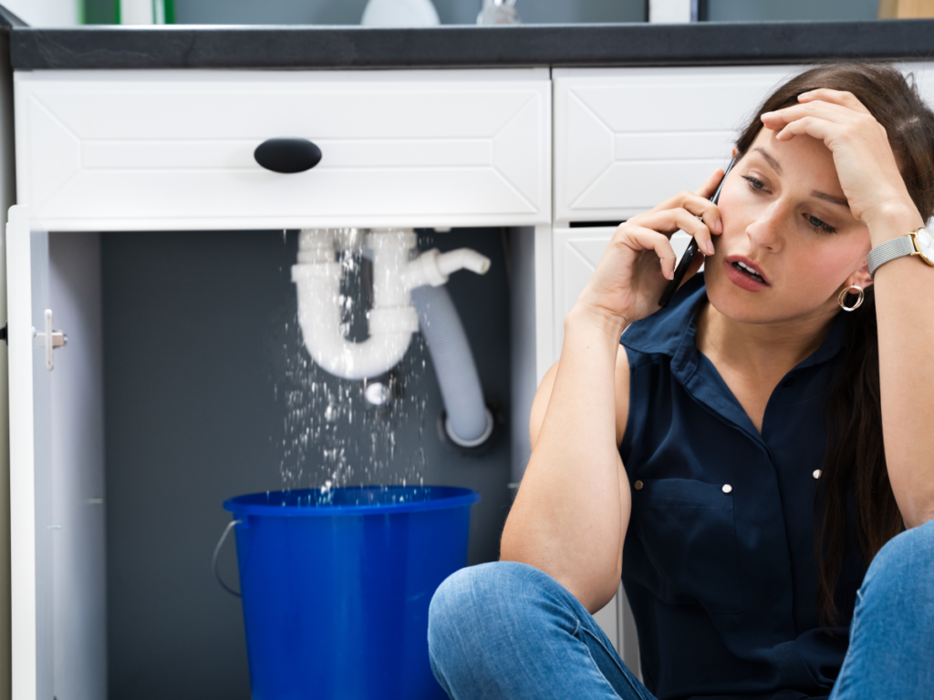 A woman sits on the floor making a phone call while high pressure water leaks from a pipe under the sink into a blue bucket.