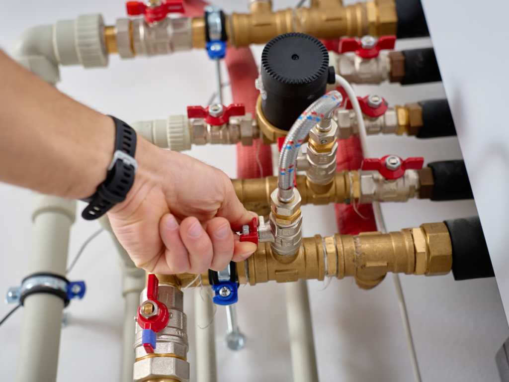A person&rsquo;s hand adjusts a valve on a network of brass water pipes, managing high pressure as gauges and red-handled shut-off valves monitor the flow.