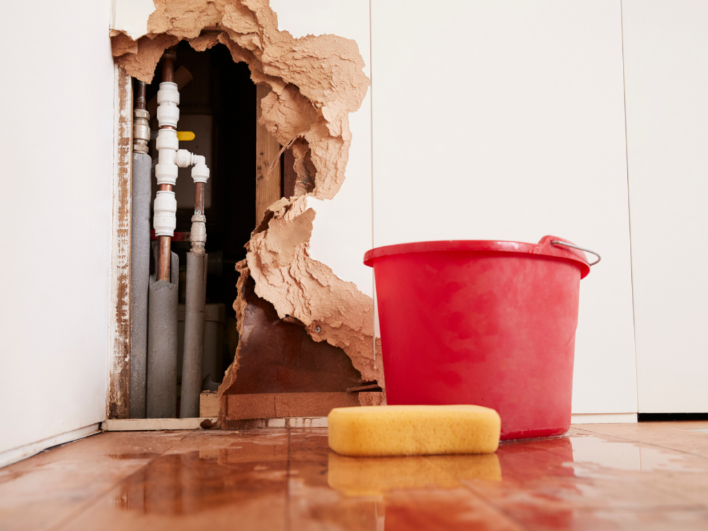 A large hole in a wall exposes high pressure plumbing pipes, with water damage on the floor, a red bucket, and a yellow sponge nearby.