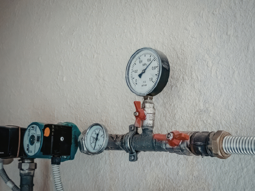 A close-up of industrial pipes with high-pressure gauges and red-handled valves mounted on a textured wall.