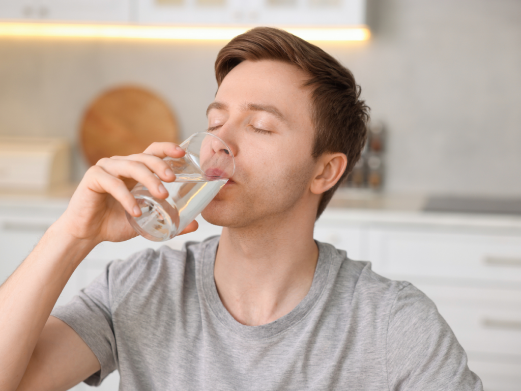 A man in a gray t-shirt is drinking a glass of water in a modern kitchen with white cabinets, unaware that rust in pipes can sometimes affect the water quality.