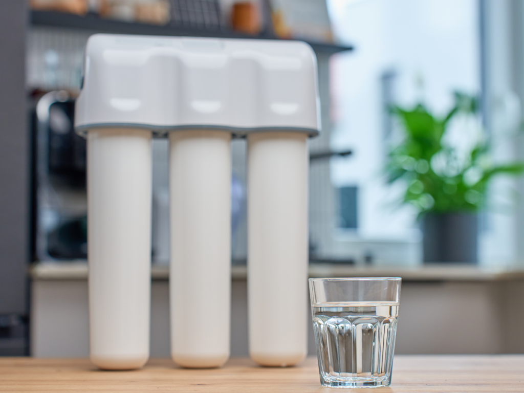 A glass of water sits on a table in front of a three-stage water filtration system, helping protect your kitchen from issues like rust in pipes.