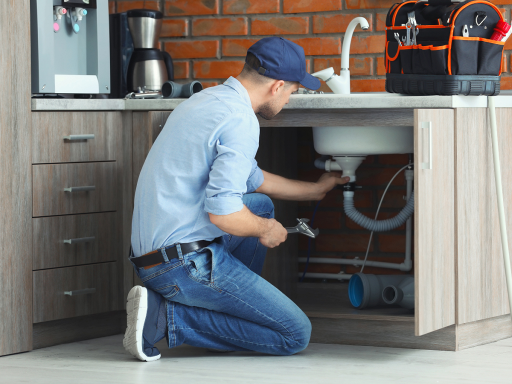 A plumber in a blue cap and shirt kneels under a kitchen sink, repairing pipes potentially affected by rust in pipes, with tools and a tool bag on the counter above.