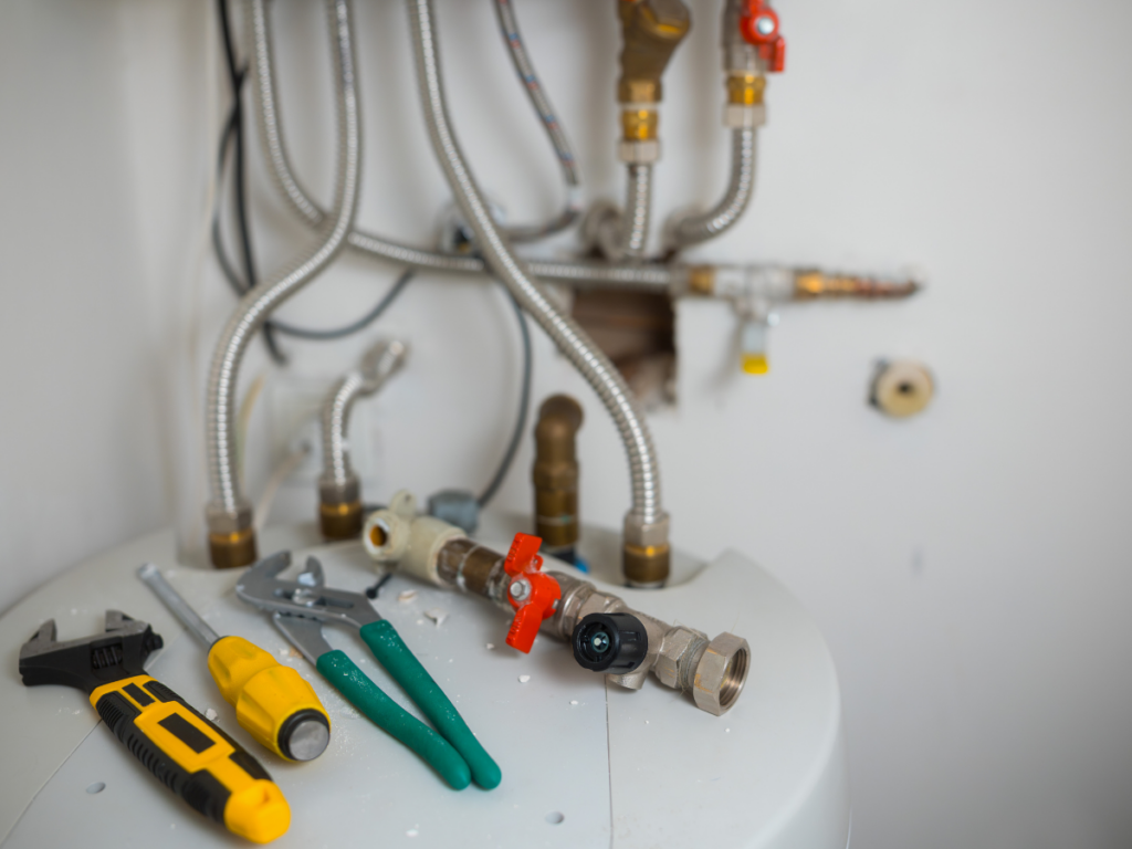 Various plumbing tools and fittings are placed on top of a water heater, with flexible metal pipes and valves visible in the background&mdash;ideal for tackling issues like rust in pipes.