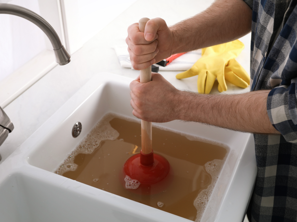 Person using a red plunger to unclog a kitchen drain clog in a sink filled with dirty water; yellow rubber gloves and a towel are nearby on the countertop.