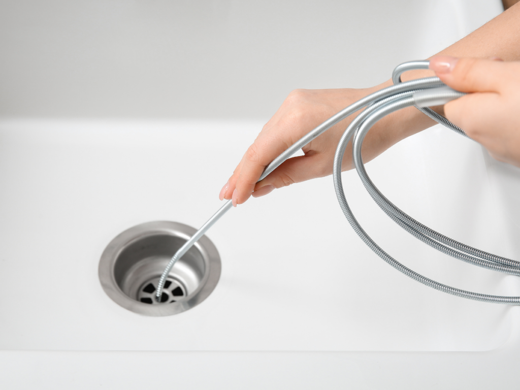 Person using a metal drain snake to clear a kitchen drain clog, with hands holding the coiled tool above a white sink.