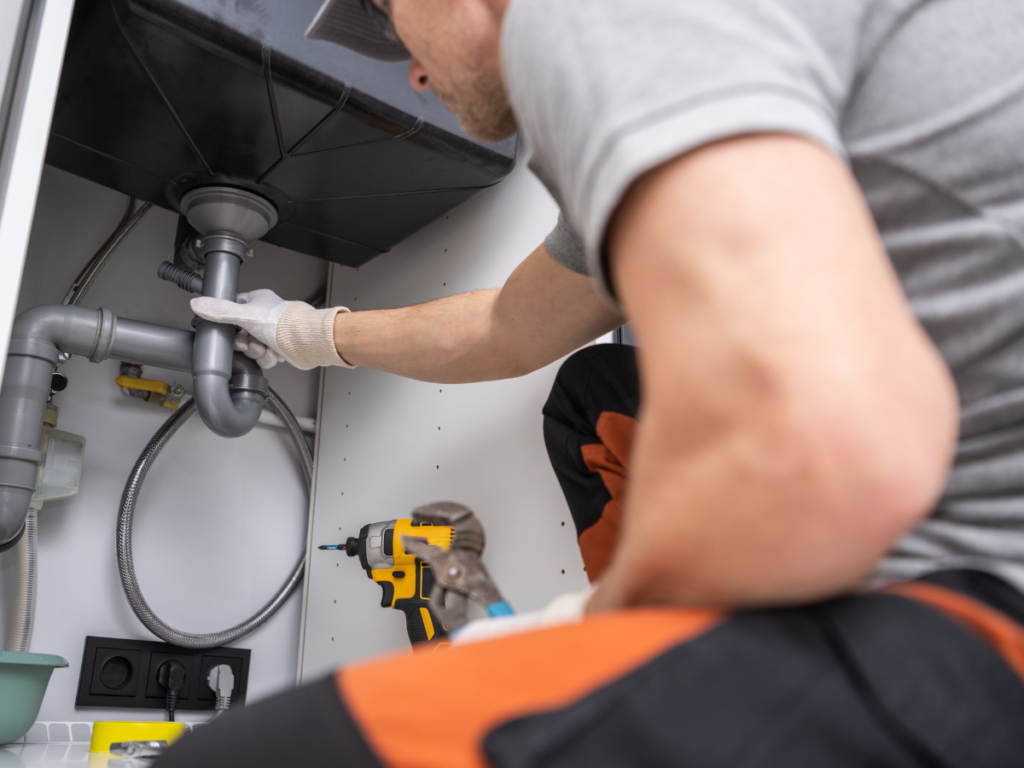 A person wearing gloves is inspecting plumbing pipes under a sink, likely addressing a kitchen drain clog, with a drill and wrench nearby.