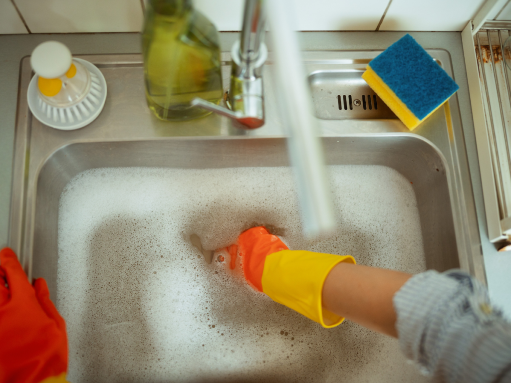 Person wearing rubber gloves washing dishes in a soapy kitchen sink, with a blue and yellow sponge and a soap dispenser nearby, ensuring the water flows freely to prevent a kitchen drain clog.