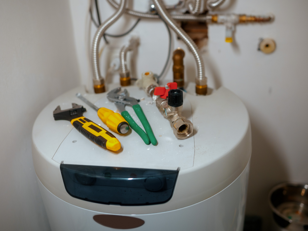 A water heater with various hand tools and a pipe fitting placed on top, showing ongoing maintenance or repair work—possibly related to addressing a kitchen drain clog.