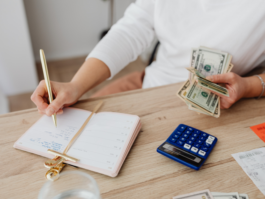 Person holding US dollar bills, writing expenses in a planner at a desk with a calculator, receipts for recent slab leak repair, and a glass of water.