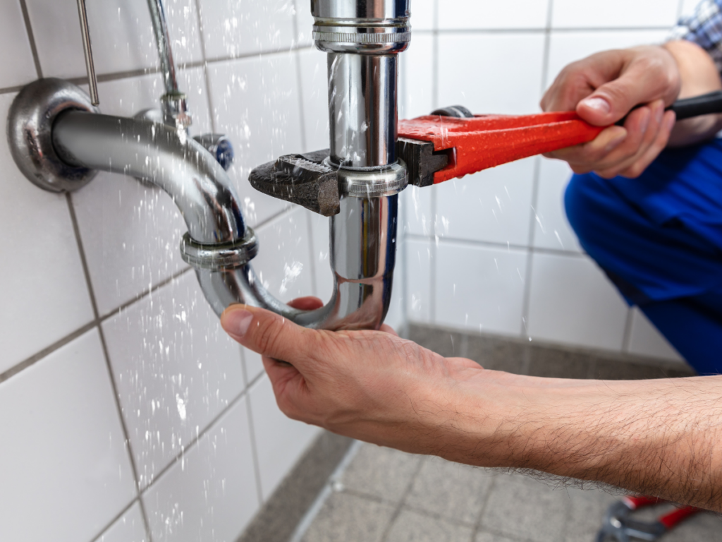 Person using a pipe wrench to fix a leaking metal pipe under a sink, addressing what appears to be a hose bib repair, with water spraying from the pipe onto the tiled wall and floor.