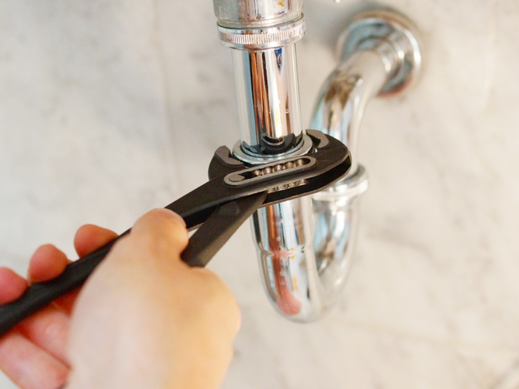 A hand uses a wrench to tighten or loosen a metal pipe under a sink, performing hose bib repair against a marble background.