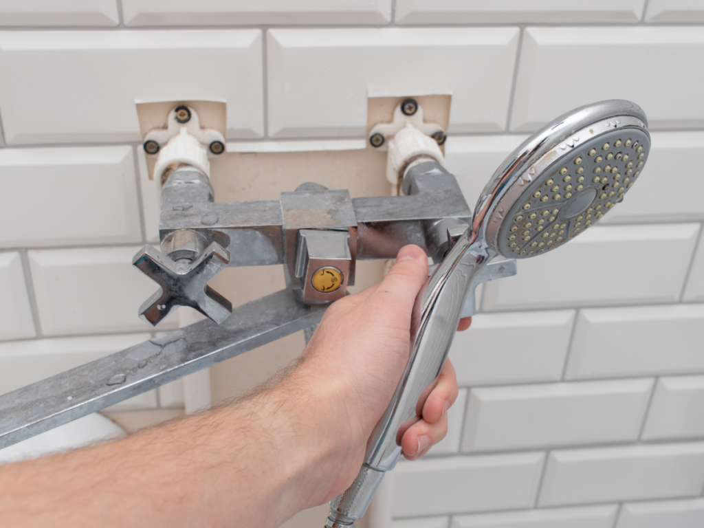 A person holds a showerhead connected to exposed plumbing fixtures on a tiled bathroom wall, appearing to perform a shower repair with two open pipe connections visible.