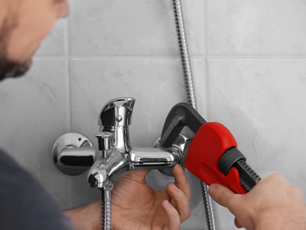 Person using a red pipe wrench to tighten or adjust a chrome shower faucet attached to a tiled bathroom wall during a shower repair.
