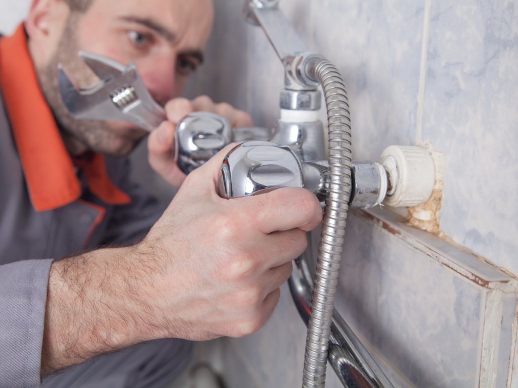 A person uses a wrench to tighten or repair a bathroom faucet with a handheld shower attachment mounted on a tiled wall, performing a shower repair.