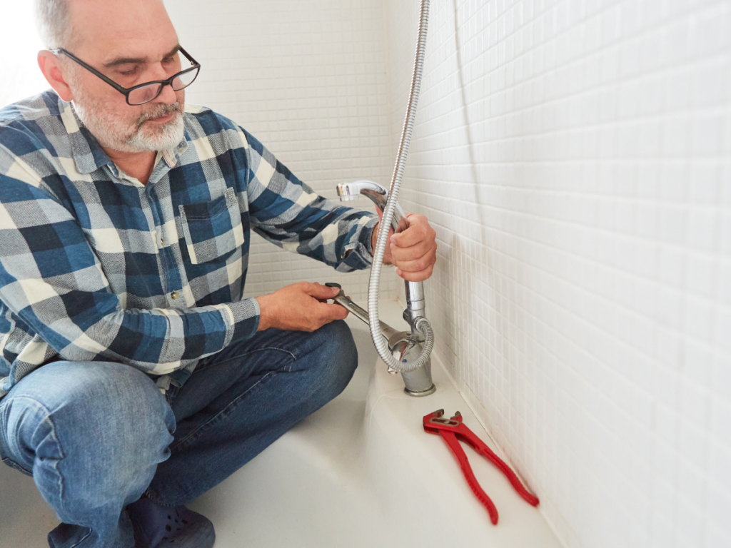 Man wearing glasses and a plaid shirt uses a wrench for shower repair on a bathtub faucet; red pliers rest on the tub beside him. A white tiled wall is in the background.