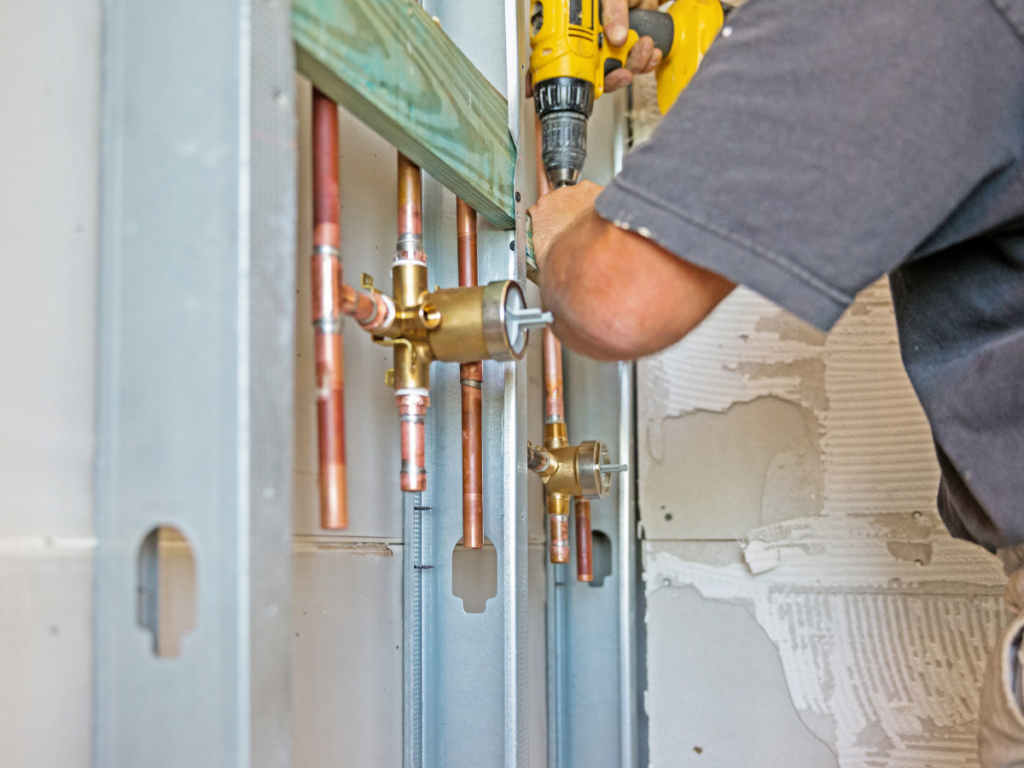 Person using a drill to install plumbing fixtures and copper pipes in a wall with exposed studs and unfinished drywall during a shower repair.