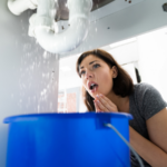 A woman looks concerned at water leaking from pipes under a sink, with a blue bucket placed below to catch the water&mdash;clearly in need of emergency plumbing assistance.