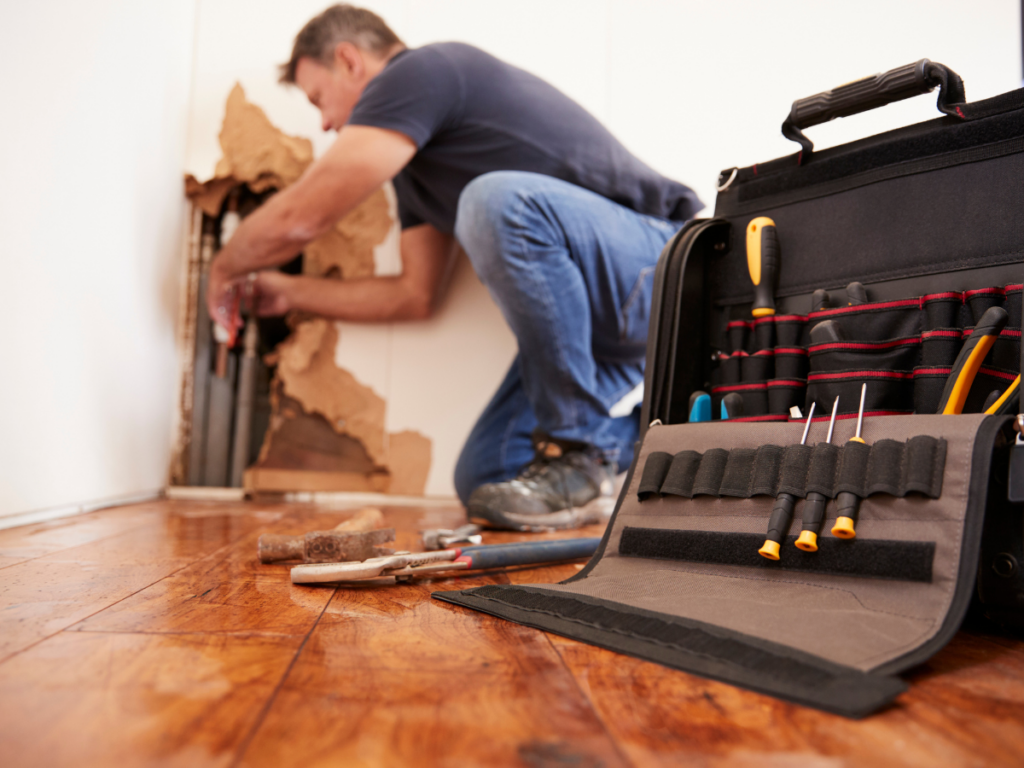 A man kneels by a damaged wall, working with pipes in what appears to be an emergency plumbing situation, while an open toolkit with various tools lies on the wooden floor nearby.
