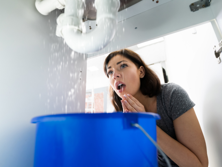 A woman looks concerned at water leaking from pipes under a sink, with a blue bucket placed below to catch the water&mdash;clearly in need of emergency plumbing assistance.