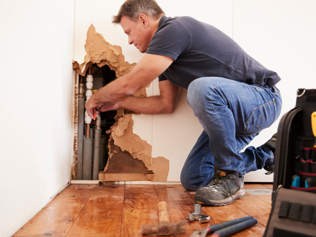 Man repairing pipes inside a wall with water damage likely caused by pinhole leaks. Tools are scattered on the wooden floor and a toolbox is nearby. The wall has a large hole exposing plumbing.