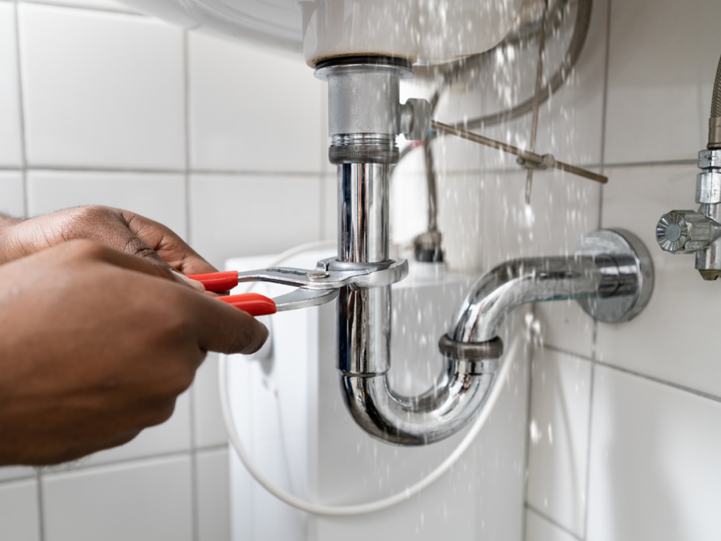 Hands using pliers to tighten a leaking metal pipe under a sink, with water spraying from the joint in a tiled bathroom&mdash;evidence of troublesome pinhole leaks compromising the plumbing.