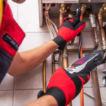 A technician wearing red gloves uses a handheld device to inspect or diagnose pipes connected to a heating or plumbing system, carefully checking for potential pinhole leaks that could cause future issues.
