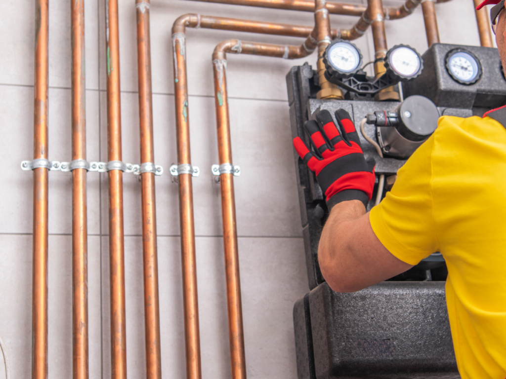 Person in yellow shirt and red gloves working on heating system controls next to several copper pipes mounted on a tiled wall, carefully inspecting for pinhole leaks.